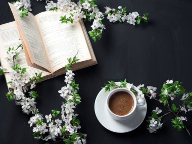 Masanın üzerinde beyaz kiraz dalları ve eski bir kitapla birlikte beyaz bir fincan espresso kahvesi. Baharın kompozisyonu Kahvaltı ve ev konforu