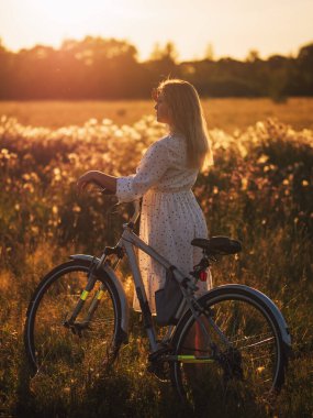 Güzel sarışın yaz günbatımının tadını çıkarıyor, yazın bisiklet sürüyor. Beyaz narin elbise