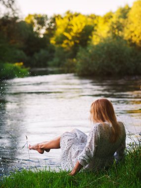 Nehir kıyısında oturan beyaz elbiseli güzel kız, güneşli bir yaz akşamı.