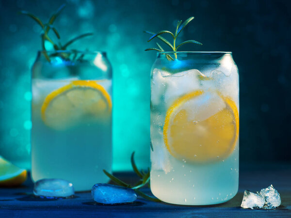 Two glasses of cold summer lemonade with lemon on the rocks and a sprig of rosemary. Colorful blue background, creative stylish glasses