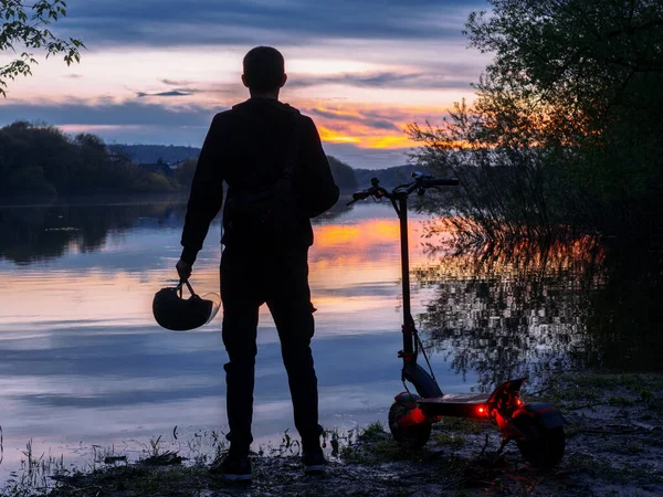 Young male rider on an electric scooter on the riverbank at sunset rear view. Colorful landscape. Modern urban transport. Freedom and love of nature, lifestyle