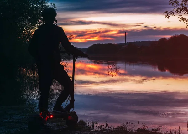 Young male rider on an electric scooter on the riverbank at sunset rear view. Colorful landscape. Modern urban transport. Freedom and love of nature, lifestyle