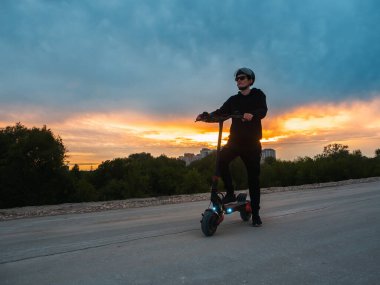 Günbatımının arka planında elektrikli scooter kullanan genç bir adam. Modern şehir ulaşımı.