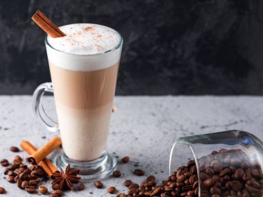 Masanın üzerinde büyük bir bardakta sütlü kahve. Kavrulmuş kahve çekirdekleri, tarçın çubukları.