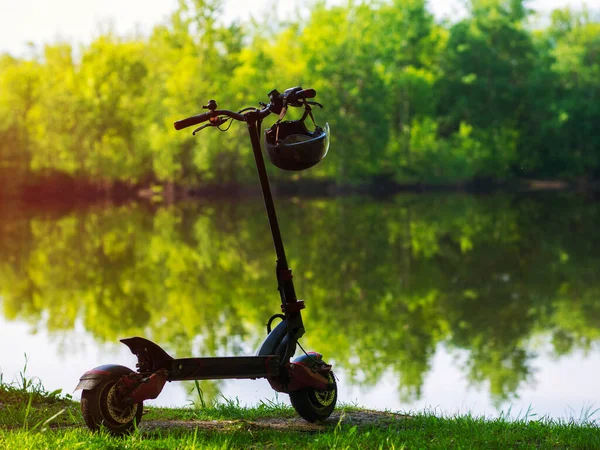 Modern electric transport. A powerful city electric scooter stands in the park on the river bank in the summer with a helmet on the steering wheel. Copy space