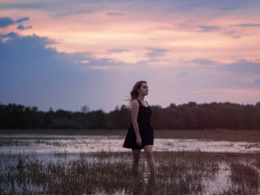 Siyah elbiseli güzel bir kadının yaz portresi. Gün batımında bir yaz günbatımının arkaplanına karşı suyun içinde durmak