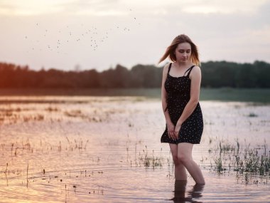 Gün batımında siyah kısa elbiseli utangaç genç bir kadın. Suda duran, gökyüzünün bir yansıması