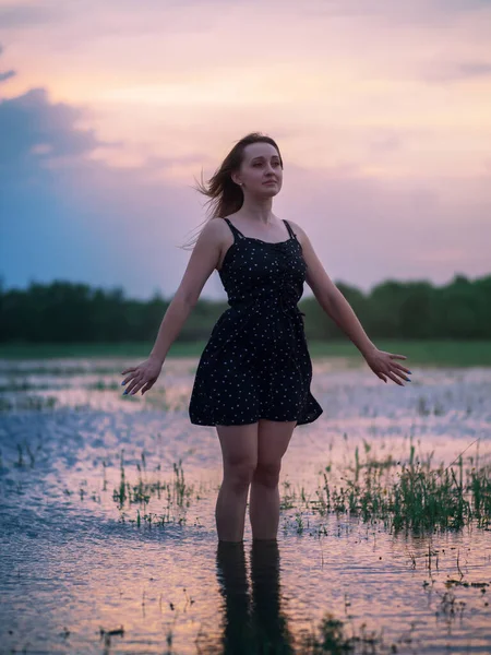 黒のドレスで美しい若い女性の夏の肖像画 夕暮れ時の夏の夕日を背景に水面に立つ — ストック写真