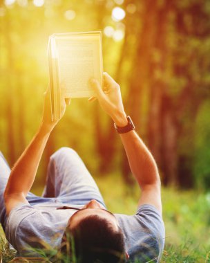 Parktaki çimlerin üzerinde uzanmış kitap okuyan genç bir adam. Açık hava eğlencesi
