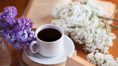 Bahar havası, ahşap bir masada bir fincan aromatik espresso kahve, çiçek açan leylak dalları ve bir kitap.