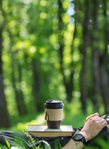 Close-up of a paper cup of coffee on a book, a man lying in the green grass in a park on a summer morning