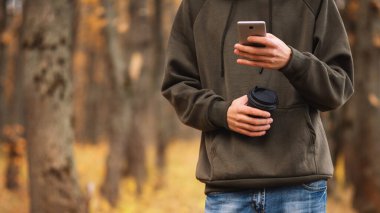 Genç bir adam sonbahar parkında yürüyor, kahve içiyor ve telefonu kullanıyor..