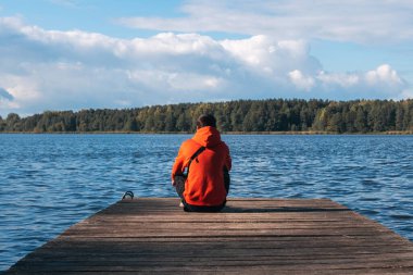 Parlak turuncu kapüşonlu genç bir adam, ahşap bir iskelede gölün arka planında. Sonbahar zamanı, seyahat ve aktif eğlence konsepti. Doğaya ve rüyaya bak.