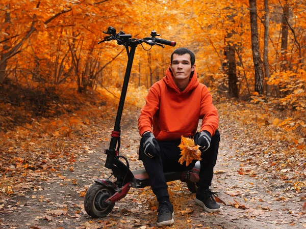 Turuncu kapüşonlu genç yakışıklı bir adam sonbahar parkında elektrikli scooter kullanıyor. Renkli mevsimlik portre, orman yolu.