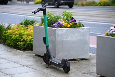 Yeşil bir elektrikli scooter şehrin kaldırımına park etmiş, çiçek tarhının yanına..