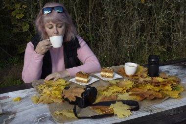 A woman in sunglasses and a pink sweater drinks coffee from a white cup at a table with desserts and dry autumn leaves. Trees and bushes form a natural fall background.