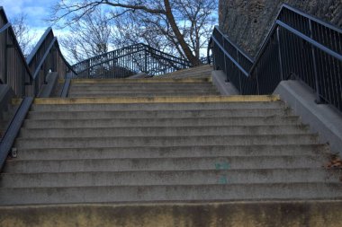 Metal parmaklıklı beton merdivenler yukarı, yapraksız ağaçlar ve arka planda taş bir duvar, açık bulutlu mavi gökyüzü, serin sonbahar havasını çağrıştırıyor..