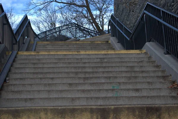 Metal parmaklıklı beton merdivenler yukarı, yapraksız ağaçlar ve arka planda taş bir duvar, açık bulutlu mavi gökyüzü, serin sonbahar havasını çağrıştırıyor..