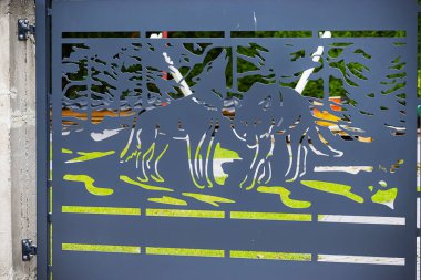 Dekoratif mavi metal kapı. Ön planda ağaç siluetleri ve iki kurt var. Kapının arkasında yeşil bitki örtüsü ve ahşap bir yapı var..