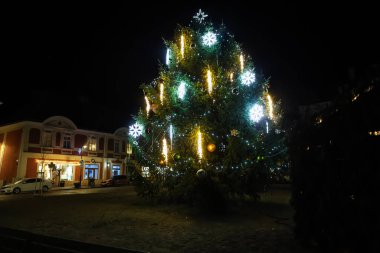 Güzel süslenmiş bir Noel ağacı bir kasaba meydanında dimdik durur. Işıkları karanlık gece gökyüzüne çarpar ve büyülü bir tatil atmosferi yaratır.