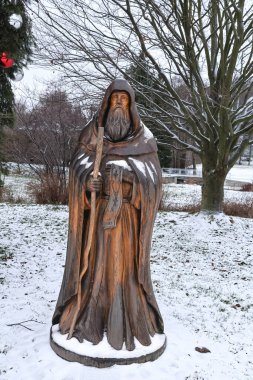 Uzun sakallı ve bastonlu cüppeli bir figürü betimleyen yıpranmış ahşap bir heykel karlı bir arazide duruyor, tarihi ve dini bir temayı ima ediyor.