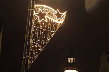 Yıldızlara benzeyen dekoratif sicim ışıkları bir sokak lambasını süsler, gece gökyüzüne karşı şenlikli bir atmosfer yaratır ve siluetli bir kilise kulesi oluşturur.