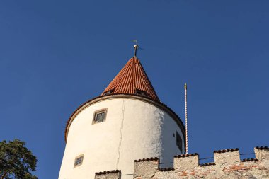 Küçük pencereleri ve kırmızı fayanslı konik çatısı olan tarihi beyaz bir kule parlak mavi gökyüzünün altında duruyor, ortaçağ mimarisi sergileniyor.