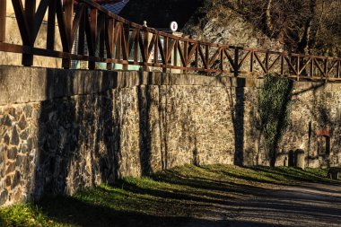 Yukarısında ahşap bir korkuluk olan taştan bir duvar. Sıcak güneş ışığıyla aydınlatılıyor. Komşu çimen yamaçlarında ve patikada dramatik gölgeler yaratıyor.