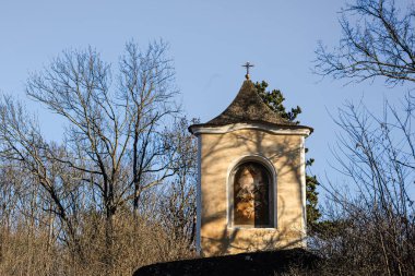 Dini bir freskle süslenmiş ve üzerinde haç olan ıssız bir yol kenarı tapınağı, parlak bir gökyüzünün altında ağaçların arasında duruyor.