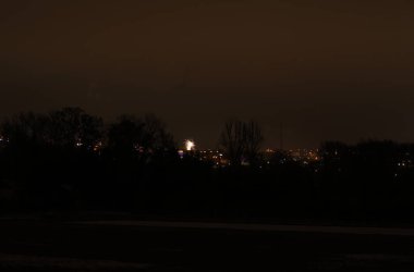 Tek bir parlak havai fişek patlaması uzak bir kasaba ve ağaçların üzerindeki karanlık gökyüzünü aydınlatıyor.