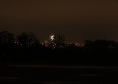 Issız bir havai fişek patlaması, uzak ve loş bir şehir manzarasının üzerindeki gece gökyüzünü aydınlatır.