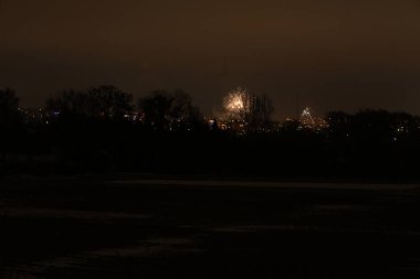 Karanlık akşam gökyüzüne karşı havai fişeklerin şenlikli gösterisi, uzak kasaba ve aşağıdaki çıplak ağaçların üzerine sıcak bir ışık saçıyor.