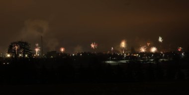 Panoramik bir görüntü, karanlık bir şehir manzarasının üzerinde patlayan havai fişek gösterisini yakalar ve şenlikli bir atmosfer yaratır.