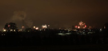 Geceleri gökyüzünde binaların ve ağaçların üzerinde sayısız havai fişeğin patladığı bir şehrin panoramik görüntüsü.