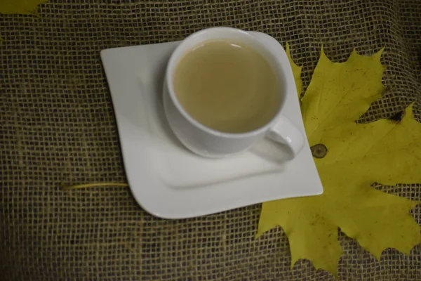 Benzersiz şekilli beyaz bir fincan üzerinde açık renkli bir içecek. Büyük sarı bir akçaağaç yaprağı, sonbahar havası yaratan, çuval bezi benzeri bir arka planda yatar..