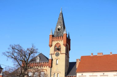 Önde gelen saat ve çan kulesinin bulunduğu eski tuğla ve taş bir kilise kulesinin detaylı görüntüsü. Parlak, bulutsuz bir gökyüzüne karşı kurulmuş.