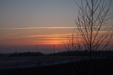 The sun dips below the horizon, casting warm hues across the sky, illuminating wind turbines in a serene countryside setting