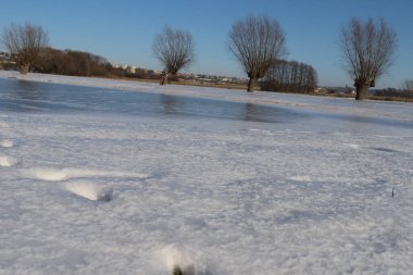 Huzurlu bir kış sahnesi, kısmen donmuş bir göletin karla kaplı zemin ve yapraksız ağaçlarla çevrili gökyüzünü yansıtmasıyla başlar.