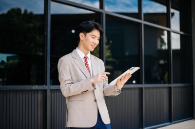 Confident Asian man with a smile standing holding notepad and tablet at out side office. big city on business center background