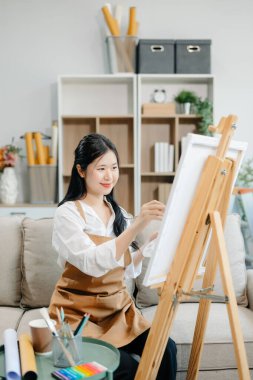 Asyalı kadın ressam sanat atölyesinde resim yapıyor, resim malzemeleri, yağlı boya tabloları, iki tuval sehpası, resim stüdyosunda veya oturma odasında fırçalı yaratıcı bir alan..
