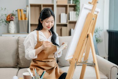 Asyalı kadın ressam sanat atölyesinde resim yapıyor, resim malzemeleri, yağlı boya tabloları, iki tuval sehpası, resim stüdyosunda veya oturma odasında fırçalı yaratıcı bir alan..
