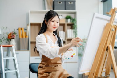 Asyalı kadın ressam sanat atölyesinde resim yapıyor, resim malzemeleri, yağlı boya tabloları, iki tuval sehpası, resim stüdyosunda veya oturma odasında fırçalı yaratıcı bir alan..