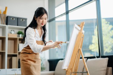 Asyalı kadın ressam sanat atölyesinde resim yapıyor, resim malzemeleri, yağlı boya tabloları, iki tuval sehpası, resim stüdyosunda veya oturma odasında fırçalı yaratıcı bir alan..