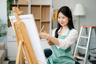 Asyalı kadın ressam sanat atölyesinde resim yapıyor, resim malzemeleri, yağlı boya tabloları, iki tuval sehpası, resim stüdyosunda veya oturma odasında fırçalı yaratıcı bir alan..