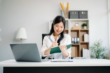 Ofiste çalışan genç iş kadını dizüstü bilgisayarı, tableti ve kâğıdı not alıyor.