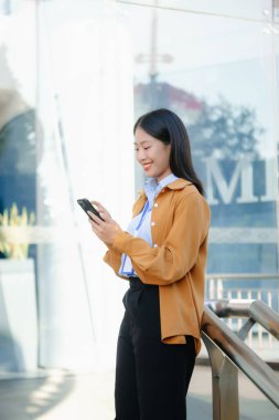 Zeki bir telefonu açık havada kullanırken gülümseyen kendine güvenen Asyalı iş kadını teknolojiyi, başarıyı ve modern profesyonel yaşam tarzını sembolize ediyor..