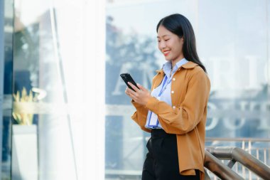 Zeki bir telefonu açık havada kullanırken gülümseyen kendine güvenen Asyalı iş kadını teknolojiyi, başarıyı ve modern profesyonel yaşam tarzını sembolize ediyor..