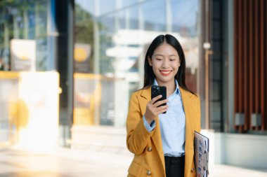 Zeki bir telefonu açık havada kullanırken gülümseyen kendine güvenen Asyalı iş kadını teknolojiyi, başarıyı ve modern profesyonel yaşam tarzını sembolize ediyor..