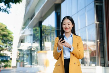 Zeki bir telefonu açık havada kullanırken gülümseyen kendine güvenen Asyalı iş kadını teknolojiyi, başarıyı ve modern profesyonel yaşam tarzını sembolize ediyor..