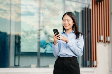 Ofisin dışında elinde not defteri ve tabletle gülümseyen kendine güvenen Asyalı kadın. İş merkezi arka planında büyük şehir